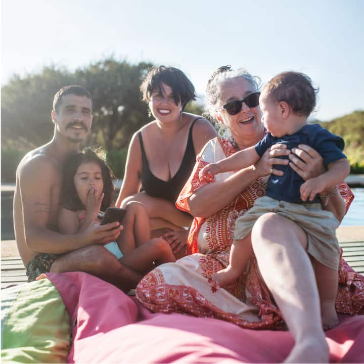 Multi-generational family with two young kids, parents, and a grandma sitting in the sun next to a pool. 