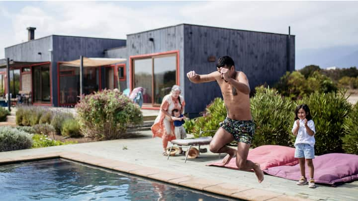 A man jumping into a pool in front of a modern, box-style house while two children and a woman look on.