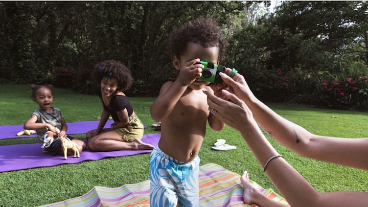 A young woman and two young children outdoors on yoga mats. One child is holding a camera and pointing it at the viewer.