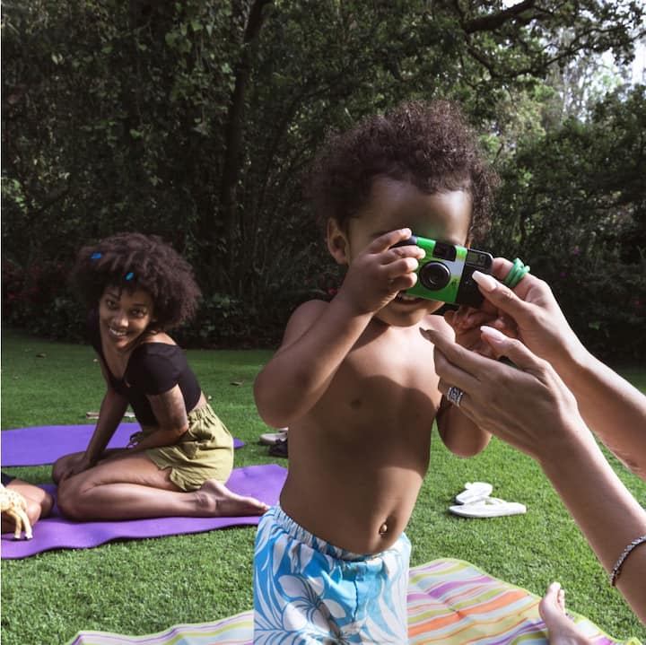 A young woman and two young children outdoors on yoga mats. One child is holding a camera and pointing it at the viewer.