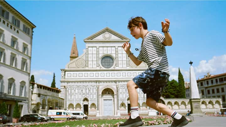 A boy in a striped shirt balancing on a step in front of an ornate European building. 