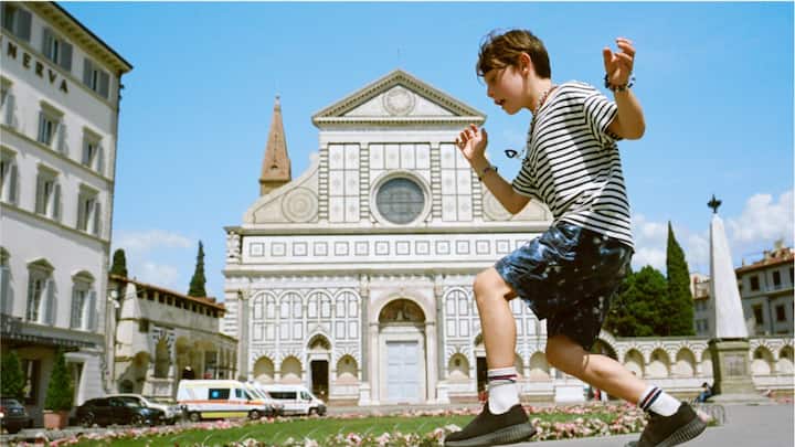 A boy in a striped shirt balancing on a step in front of an ornate European building. 
