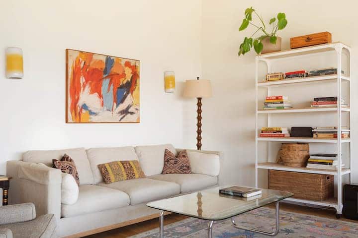 Living room with a beige couch glass coffee table, caustic abstract painting, and a book shelf with small stacks of books