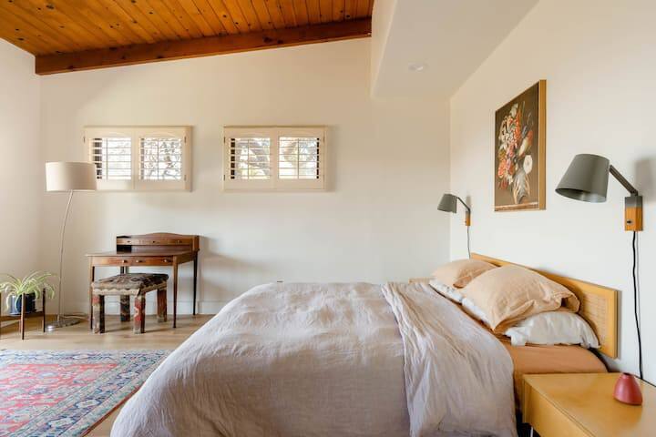 A Bedroom: the bed has two pillows on each side, and a lamp above each side. The ceiling has wooden panels. The small green plant and patterned rug make the room look cozy.