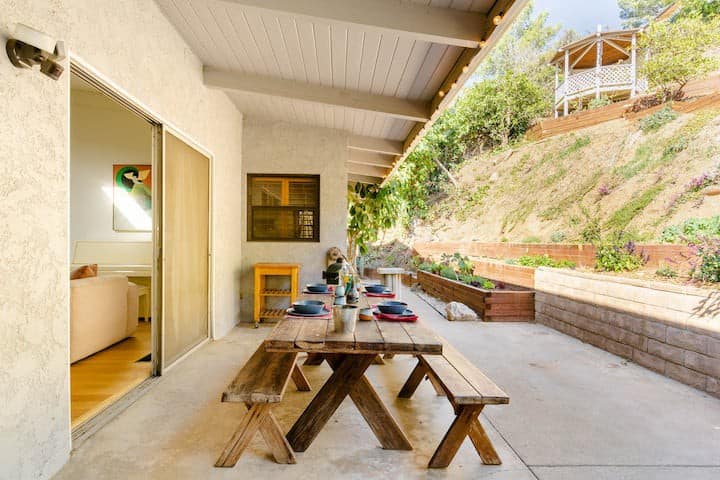 Outdoor patio featuring a wooden picnic table with benches. On the table is table settings with plates and bowls. The backyard is lit by the afternoon sun.