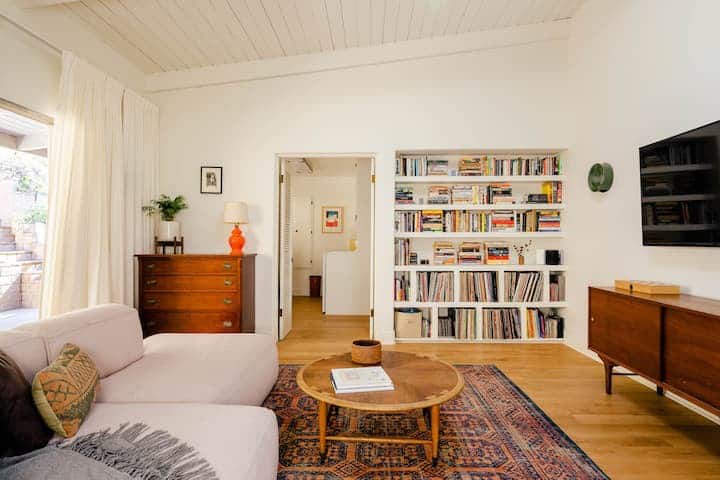 Uma sala de estar aconchegante: um lindo tapete e um sofá branco novo e fofo. Uma mesa de madeira em frente a uma estante embutida cheia de livros e discos de vinil.