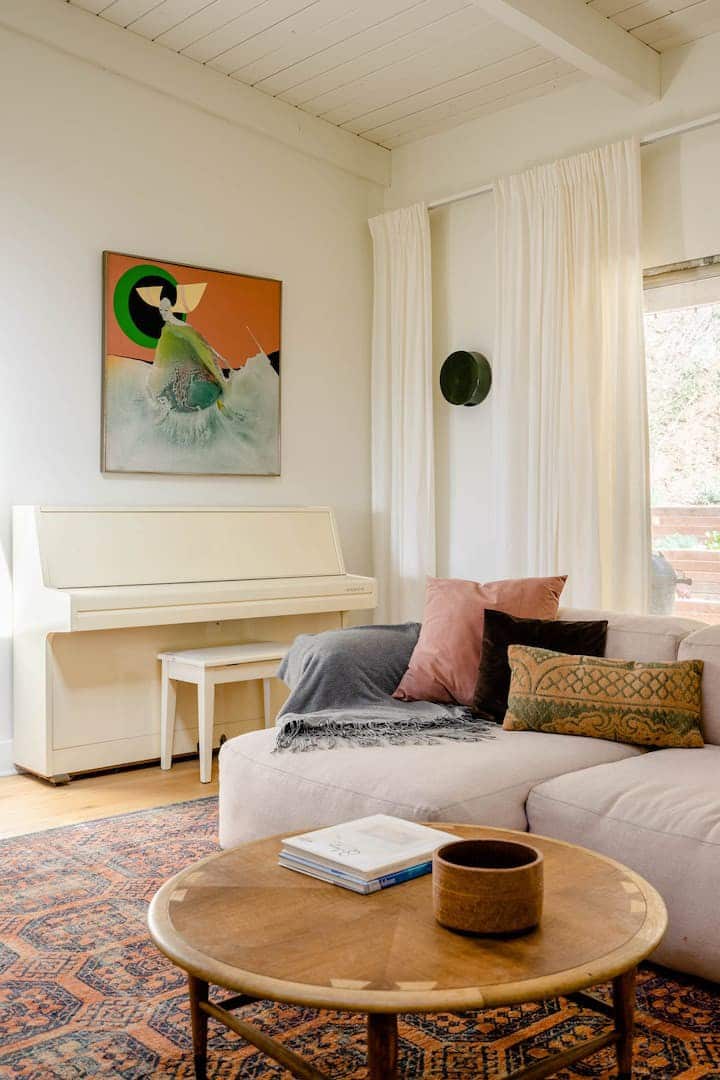 The corner of the living room features a small white upright piano. Above it sits a painting with orange and green tones. The blush colored couch has accent pillows of many colors, and a grey-lavender throw blanket.