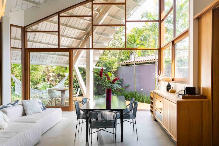 A Brazilian living room with a lot of beautiful light is coming through a wall made entirely of glass, and in front is a dining table with fresh tropical flowers.