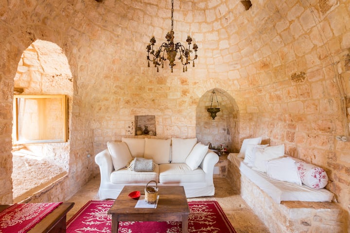 Beroemde Italiaanse trullo in een koepelachtig bouwwerk van warme stenen. Een woonkamer met een witte bank, een rood tapijt en een vorstelijke lamp erboven.