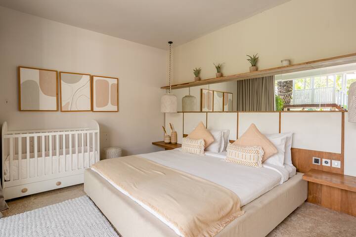 A bedroom with a queen size bed, and a white baby crib. The tones in the room are creamy and beige. It is a nicely style room.