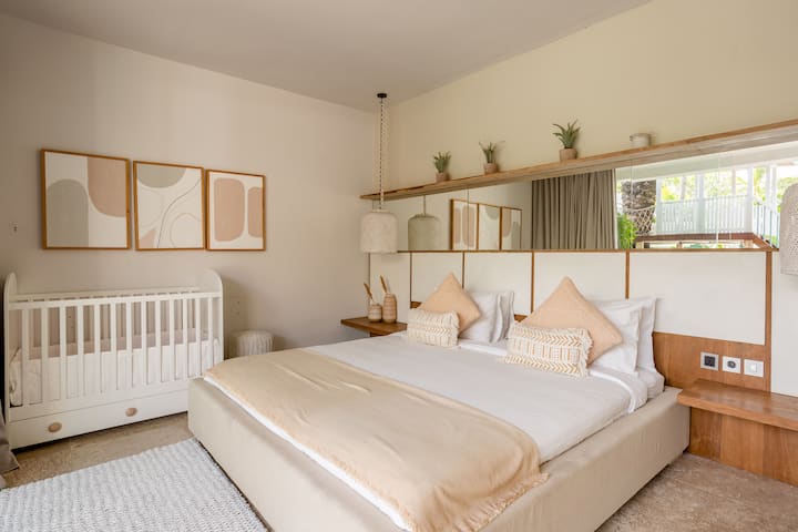 Un dormitorio con una cama queen y una cuna de color blanco. La habitación tiene tonos pastel y beige y está decorada con gusto.