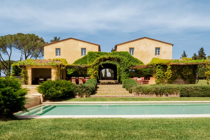 Ein Haus im toskanischen Stil mit Bögen, die mit Weinreben bewachsen sind, hat einen großen, luxuriösen Pool, der im Vordergrund sichtbar ist. Es ist ein wunderschöner sonniger Tag mit blauem Himmel und zarten Wolken.