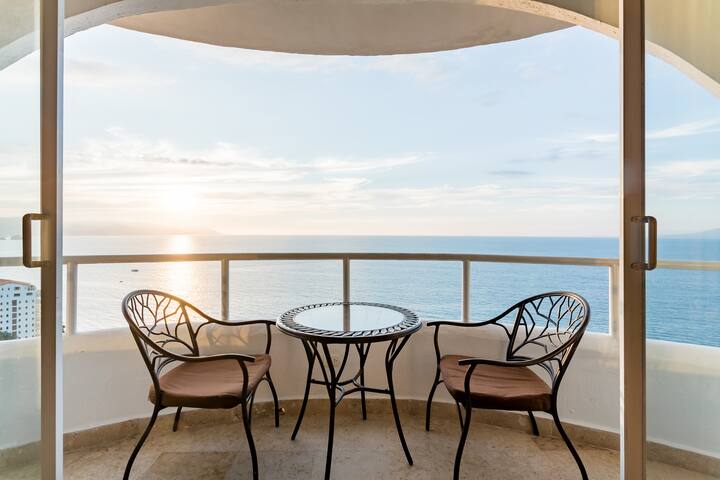 Doors open on a balcony with a small table and a chair on either side. The balcony looks out on a beautiful ocean at golden hour. 