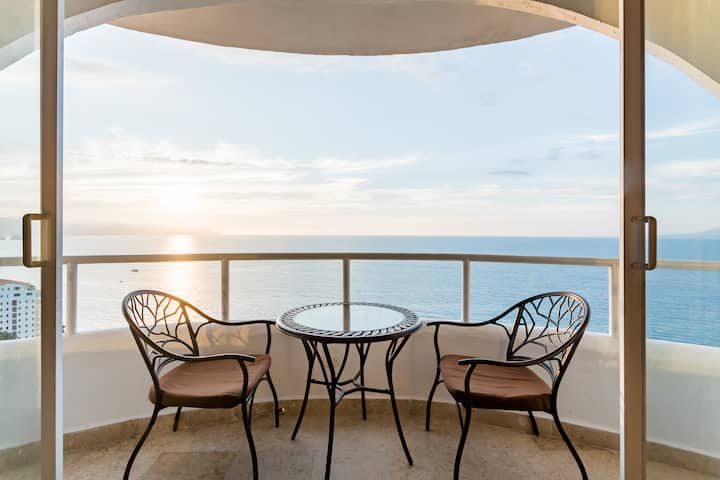Doors open on a balcony with a small table and a chair on either side. The balcony looks out on a beautiful ocean at golden hour. 