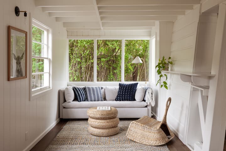 A small living room, with a couch from wall to wall. The room is painted white, and has a wicker table and wicker chair on top of a rug with similar earthy tones. The window behind the couch has a beautiful collection of trees, creating a nice green ambiance.