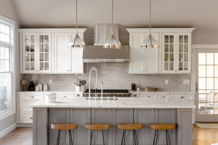 A beautiful large kitchen featuring a large island with 4 stools in front. 3 lamps hang down from the ceiling above the island. The kitchen is made up of grey and beige tones.