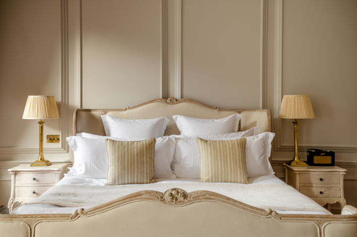 A bedroom with fluffy pillows. The room is very symmetrical with side tables and lamps on each side of the bed. The walls and bed frame are a creamy beige color, and the beds sheets are bright white.