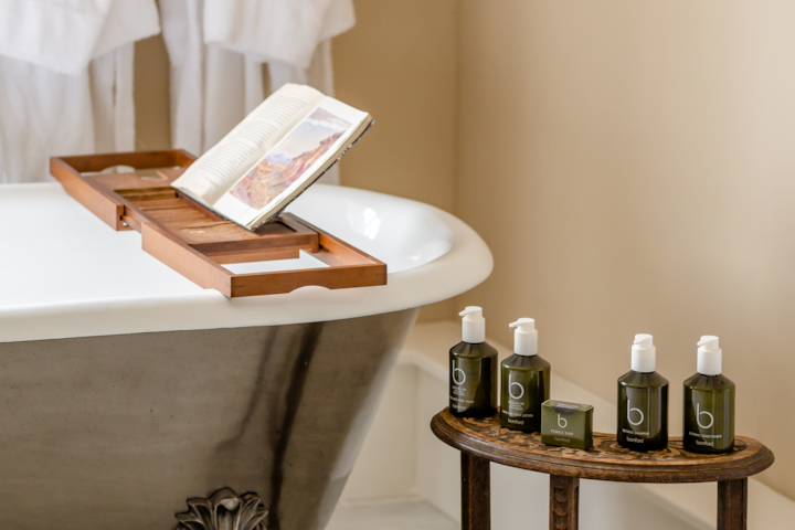 A close up of a clawfoot bathtub. On a stool next to the tub sits small bottles of bespoke soap, shampoo, and conditioner. Two terry cloth bathrobes hang from a hook in the background.