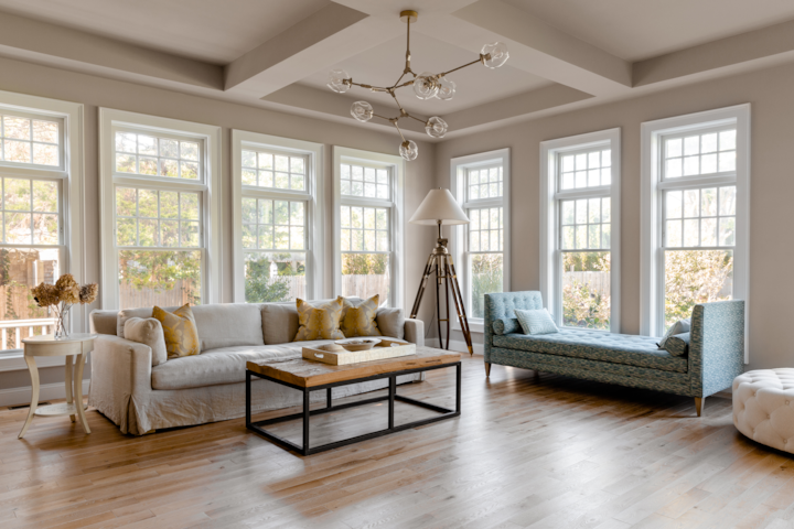 A living room with a beige couch, and a teal patterned sunbed. There are 7 long and large windows (almost floor to ceiling). A lamp on a wooden tripod sits in the corner. The furniture sits on a wooden floor. The light is soft.