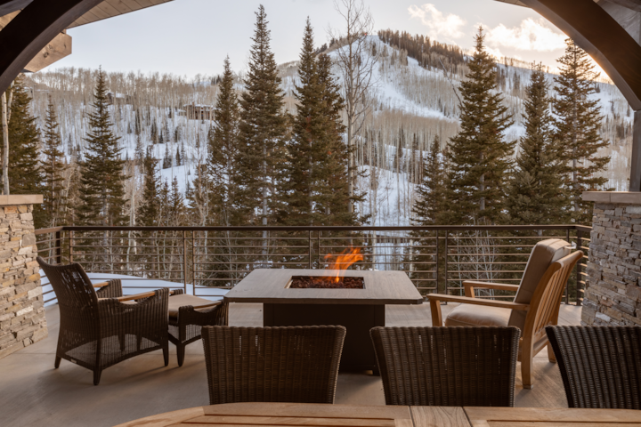A balcony featuring outdoor furniture and a lit fireplace overlook a beautiful mountain filled with snow. The image is framed by an arching beam. It's is late afternoon light. It's a very inviting and cozy balcony.