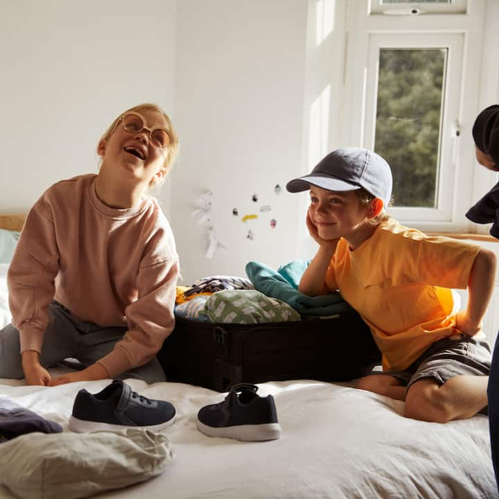 Two little kids playfully pack a suitcase on a bed. 