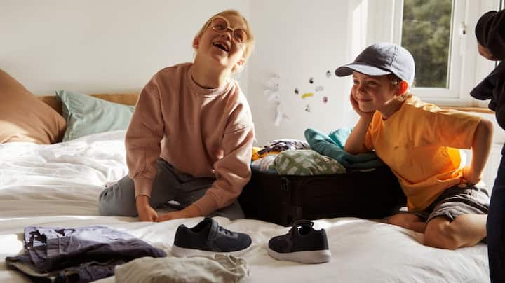 Two little kids playfully pack a suitcase on a bed. 