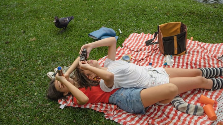 Two children at a picnic look up at the sky through binoculars. 