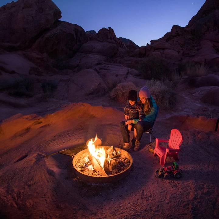 A little boy sits on his mom’s lap in front of a desert fire pit at night. 