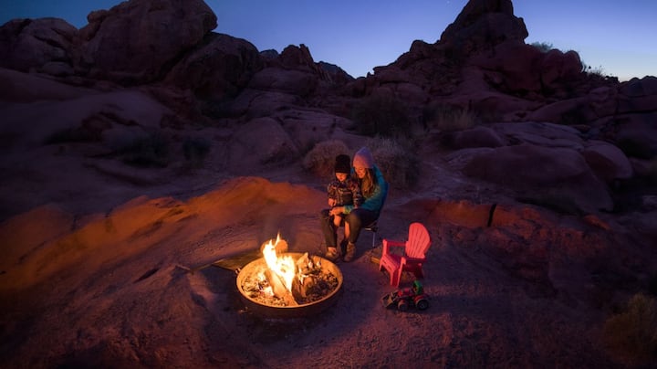 A little boy sits on his mom’s lap in front of a desert fire pit at night. 