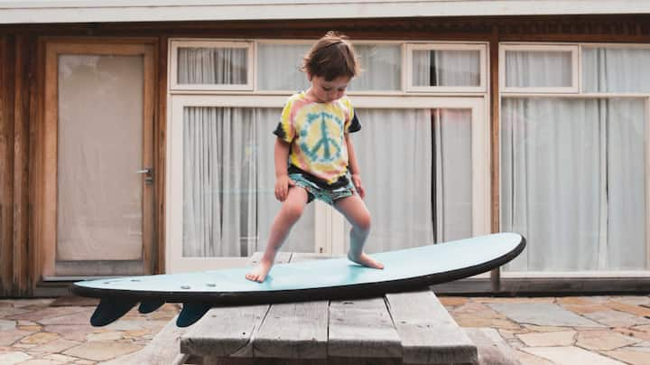 A toddler in a peace sign t-shirt practices standing on a surfboard.