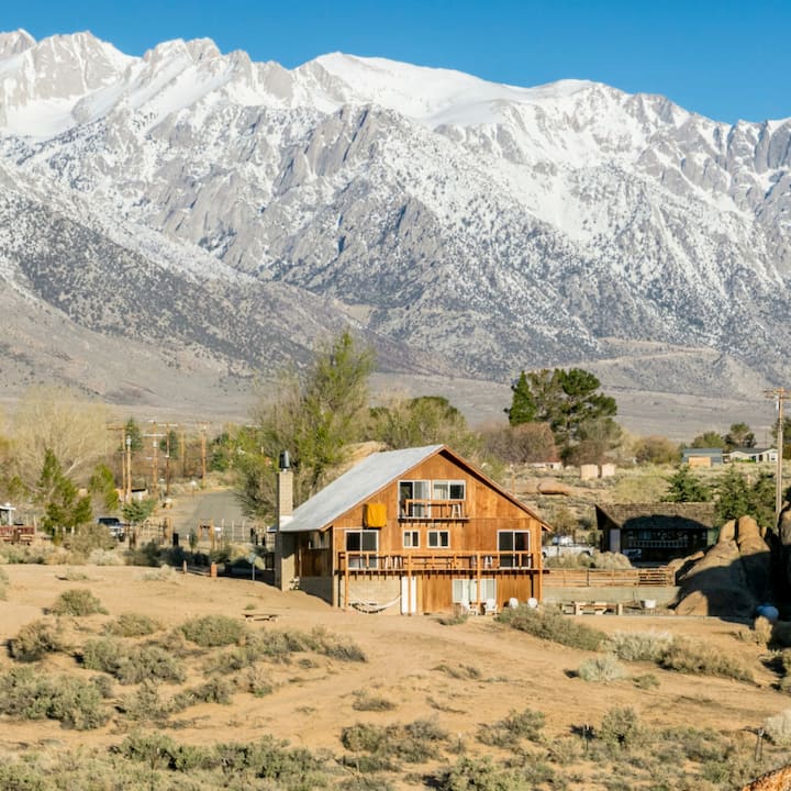 A sun-drenched cabin sits at the base of snow-topped mountains.