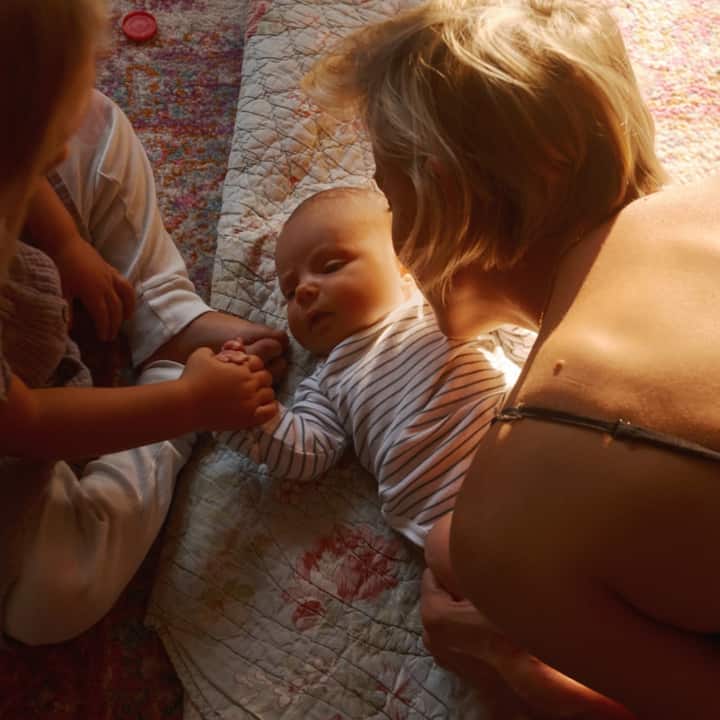 A baby is being held by her mom while holding hands with her older sister.
