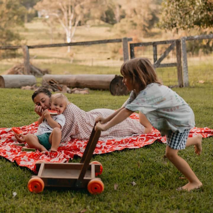 A toddler pushes a play cart around the park as her little brother looks on. 