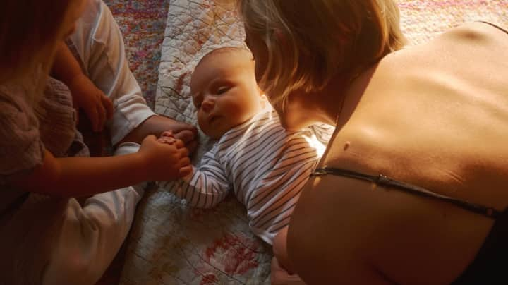 A baby is being held by her mom while holding hands with her older sister.