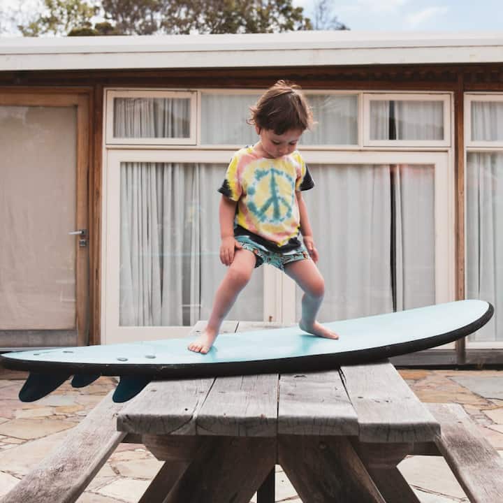 A toddler in a peace sign t-shirt practices standing on a surfboard.