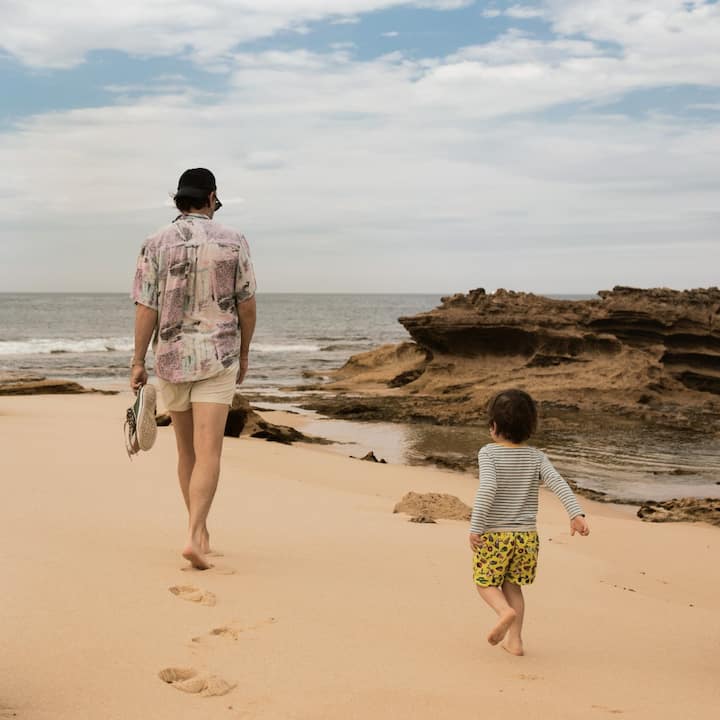 A father and son stroll barefoot around a beach on an overcast day. 