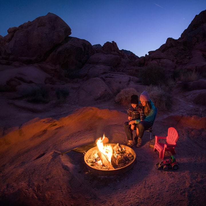 A little boy sits on his mom’s lap in front of a desert fire pit at night. 