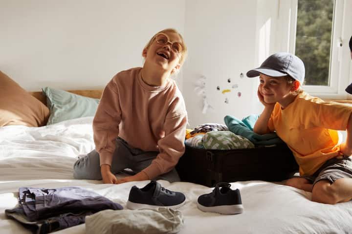 Two little kids playfully pack a suitcase on a bed.