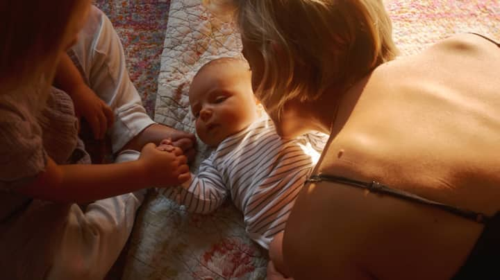 A baby is being held by her mom while holding hands with her older sister.