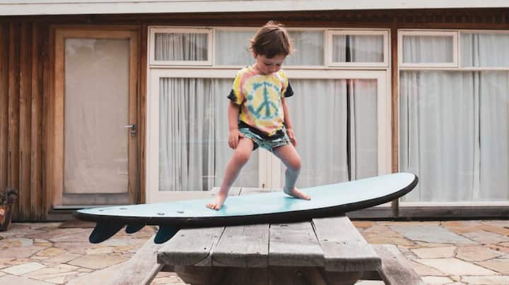 A toddler in a peace sign t-shirt practices standing on a surfboard. 