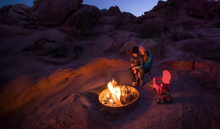 A little boy sits on his mom’s lap in front of a desert fire pit at night. 