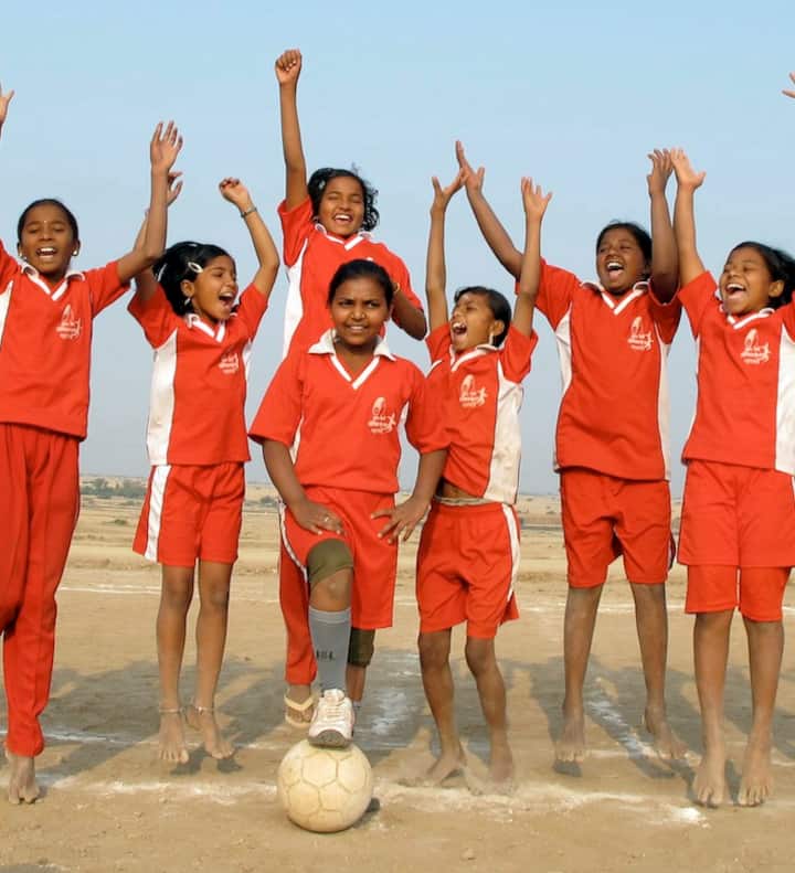 Children wearing red and white uniforms jump in the air smiling, while one rests a foot on a soccer ball.