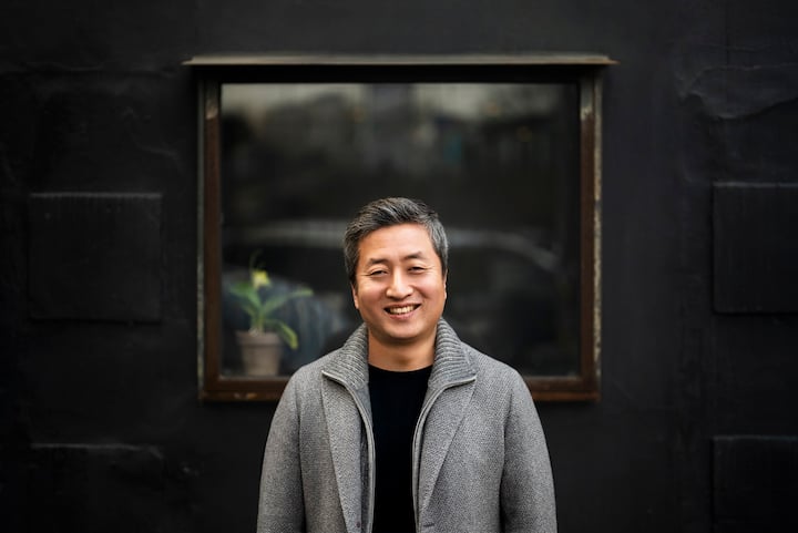 A person wearing a gray sweater blazer smiles standing in front of a window and dark wall.