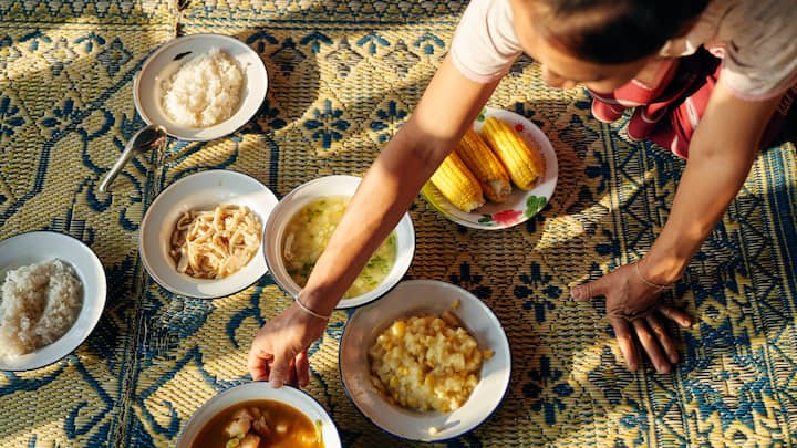 Una persona está de rodillas reclinada sobre varios tazones blancos con comida que se han colocado sobre una alfombra de color tostado y azul.