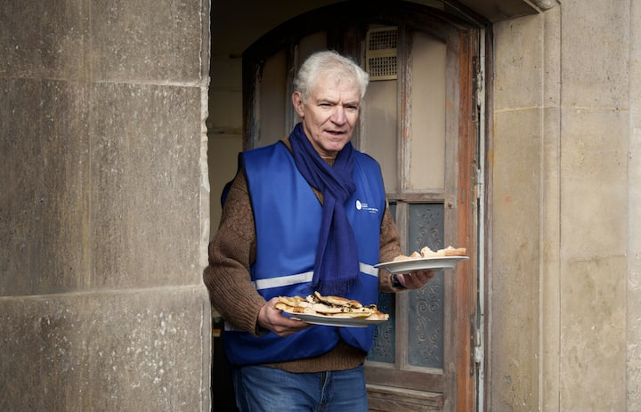 Uma pessoa com lenço e colete azul sai de um prédio de pedra carregando dois pratos de comida.