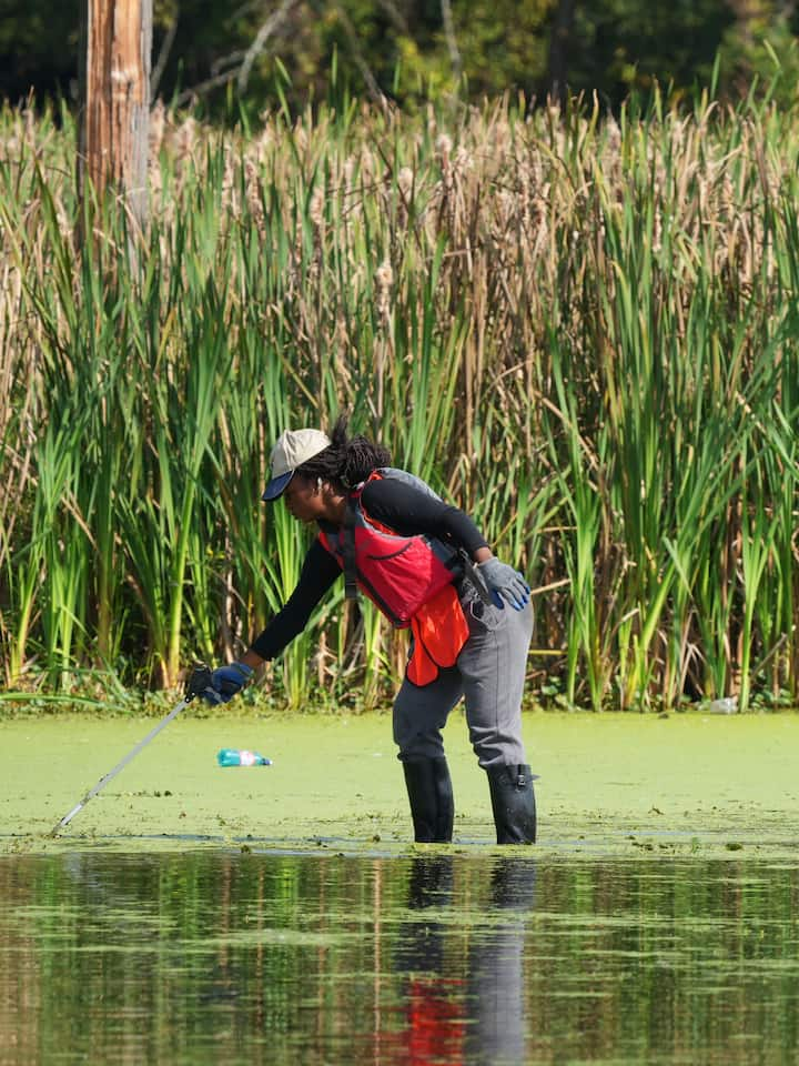 Una persona con botas de agua altas está de pie en una superficie de aguas poco profundas junto a una pequeño barco azul.