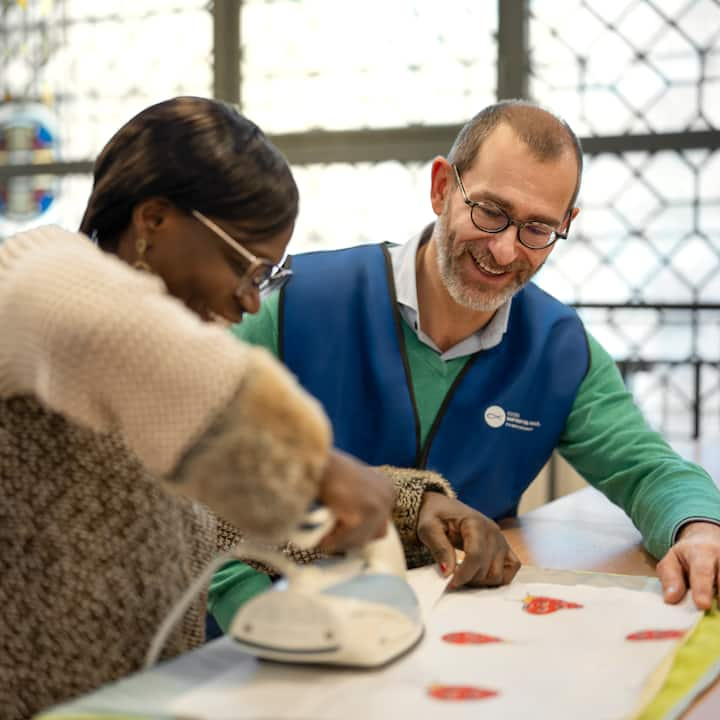 A seated person irons a piece of fabric on a table while another seated person looks on.