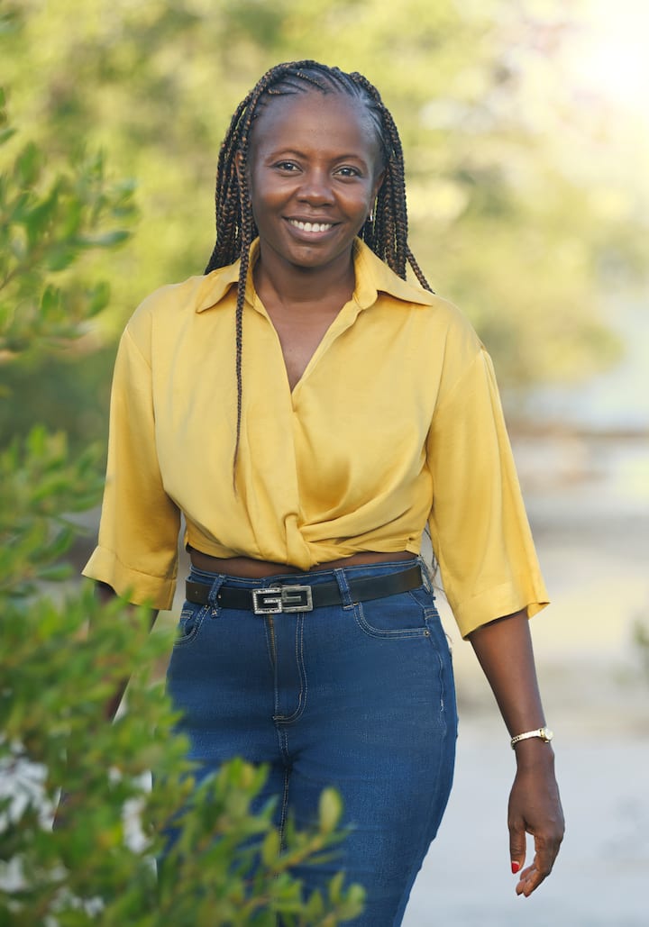 A person with long braids and a yellow blouse smiles in front of a grove of trees.