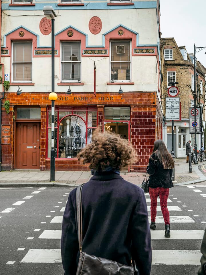 Três pessoas usando jaquetas e bolsas de ombro atravessam uma rua repleta de prédios de tijolos.