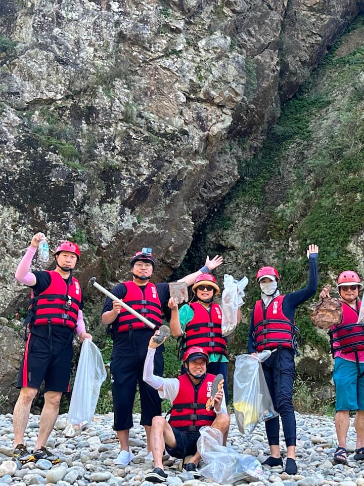 Eine Gruppe von Menschen mit roten Westen und Müllsäcken steht auf felsigem Untergrund während einer Aufräumaktion an einem Fluss.